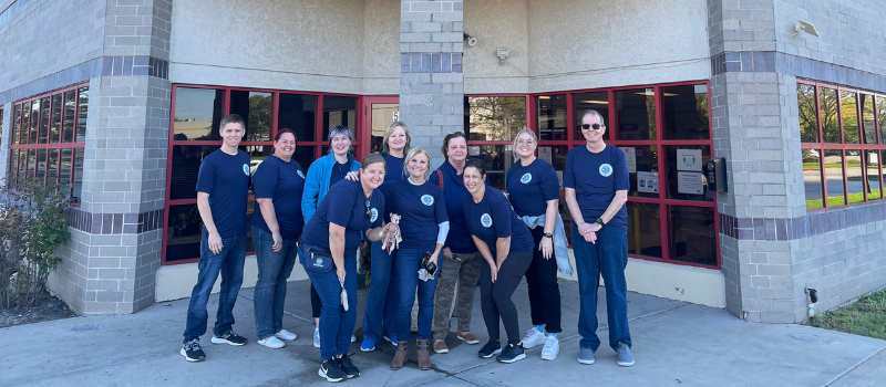 A group of eleven people wearing matching blue shirts poses for a photo outside a building with large red-framed windows.