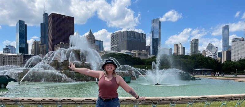 Person standing in front of a large fountain with arms outstretched, skyscrapers and a blue sky with clouds visible in the background.