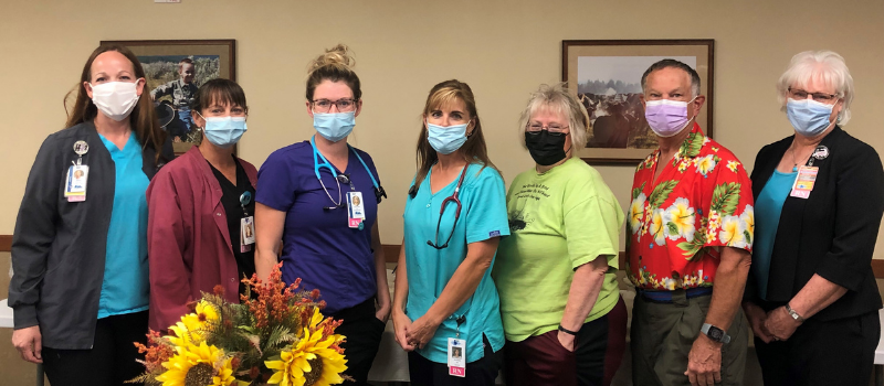 Seven adults wearing face masks and work badges stand in a row indoors, with sunflowers in the foreground and framed pictures on the wall behind them.