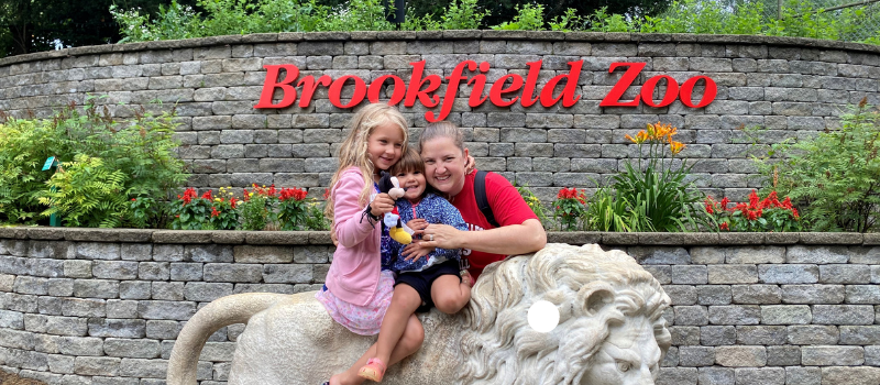 A woman and two children sit on a white lion statue in front of a brick wall with a red "Brookfield Zoo" sign and flowers.