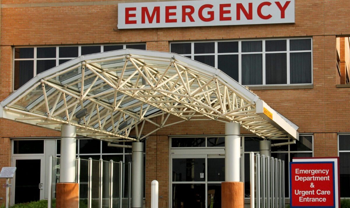 Entrance to a hospital emergency department with a large "EMERGENCY" sign above the door and a covered walkway leading to the entrance.