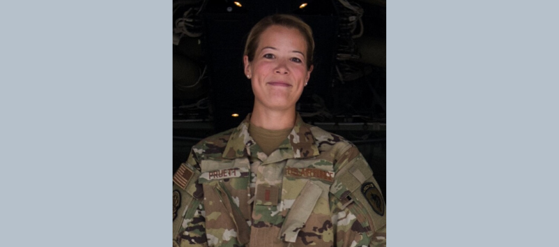 A person wearing a U.S. military camouflage uniform stands in front of a dark background, facing the camera and smiling.