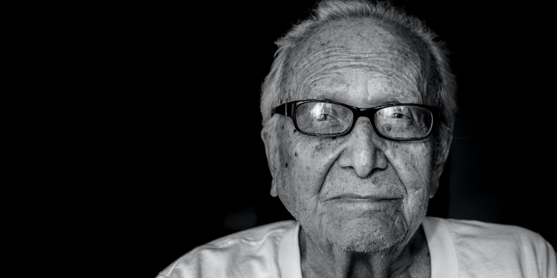 Black and white close-up portrait of an elderly man wearing glasses and a white shirt, looking slightly to the side with a neutral expression.