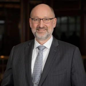 A middle-aged man with glasses, a beard, and a bald head is wearing a dark pinstripe suit, a patterned tie, and is smiling at the camera.