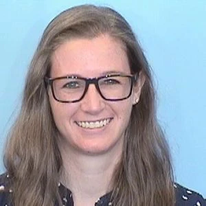 A woman with long brown hair and glasses smiles at the camera against a plain light blue background.
