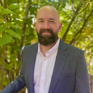 A man with a beard and bald head wearing a gray suit jacket and white shirt stands outdoors in front of green leafy trees.