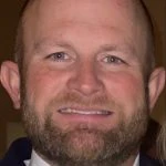 A man with a full beard and short hair is facing the camera and smiling slightly.