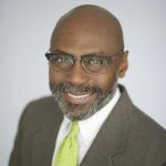 A man with a gray beard and glasses wearing a brown suit, white shirt, and light green tie, smiling at the camera against a plain light background.
