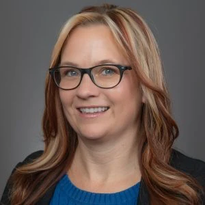 Woman with long, light brown hair with blonde highlights, wearing glasses, a blue top, and a black blazer, smiling at the camera against a plain gray background.