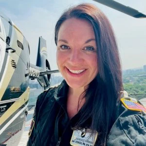 A woman in a pilot uniform stands in front of a helicopter, smiling at the camera. The helicopter has an American flag patch and cityscape is visible in the background.