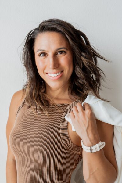 Woman with shoulder-length brown hair wearing a sleeveless ribbed tan top, holding a white jacket over her shoulder, smiling at the camera—one of the inspiring BCEN conference speakers against a plain background.