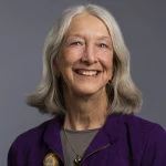 An older woman with gray hair and a purple jacket smiles at the camera against a plain gray background.
