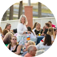 A woman stands and smiles among a group of people seated outdoors at tables, some with drinks and conference name badges, during a daytime emergency nursing conference sponsored by an exhibitor.