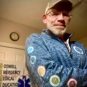 Man wearing a blue zip-up jacket with patch badges, glasses, and a cap stands indoors near a wall sign reading "Roswell Emergency Medical Education." A clock hangs above the sign.