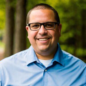 A man wearing glasses and a light blue collared shirt smiles outdoors with trees and greenery in the background, capturing the approachable energy of BCEN conference speakers.