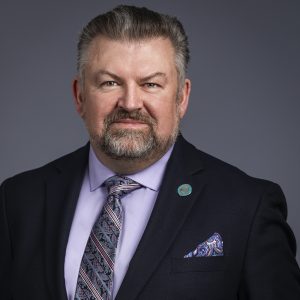 A middle-aged man with short gray hair and a beard, wearing a dark suit, patterned tie, and lavender shirt, poses against a plain gray background—resembling one of the distinguished BCEN conference speakers.
