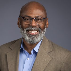 A man with a bald head, glasses, and a gray beard smiles while wearing a brown suit jacket and blue shirt against a plain gray background, embodying the professionalism of BCEN conference speakers.
