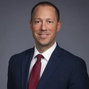 A man in a navy blue suit, white shirt, and red tie smiles at the camera against a plain gray background.