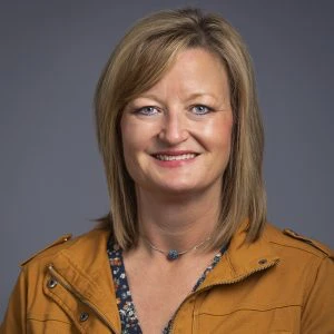 A woman with blonde hair, wearing a mustard yellow jacket over a floral top, smiles at the camera against a plain gray background.