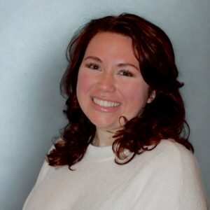 A woman with wavy brown hair, wearing a white sweater, smiles at the camera against a plain light blue background, embodying the confident and approachable presence of BCEN conference speakers.