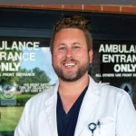 A man in medical scrubs and a white coat stands in front of a hospital entrance marked "Ambulance Entrance Only.