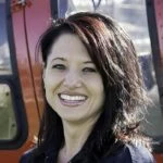 A woman with dark hair smiles in front of a red vehicle, possibly a helicopter.