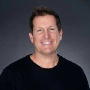 Smiling man with short brown hair wearing a black shirt, shown against a plain dark background.