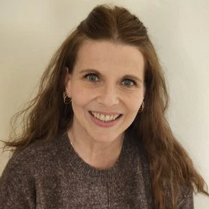 A woman with long brown hair, wearing a brown sweater and small hoop earrings, smiles at the camera in front of a plain light-colored wall.