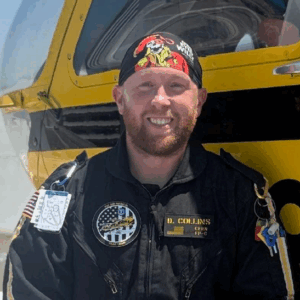 A man in a black flight suit with patches and a pirate-themed bandana, reminiscent of BCEN conference speakers, sits in front of a yellow and black aircraft, smiling at the camera.