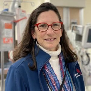 Woman with long brown hair and red glasses, wearing a blue jacket and colorful scarf, stands in a medical setting with monitors and equipment in the background.