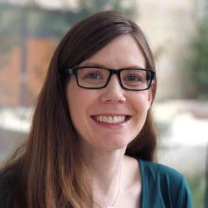 Woman with long brown hair and glasses smiling, wearing a teal top, seated indoors with a blurred background.