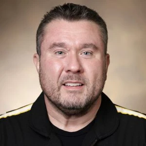 A man with short brown hair and a beard is wearing a black jacket with yellow stripes, posing in front of a neutral brown background.