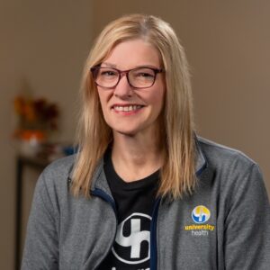 A woman with straight blond hair and glasses, wearing a gray University Health jacket, smiles at the camera in an indoor setting, representing BCEN conference speakers.