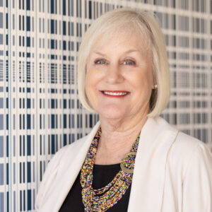 A smiling older woman with short white hair, one of the BCEN conference speakers, wears a white blazer, black top, and colorful beaded necklace while standing in front of a patterned wall.