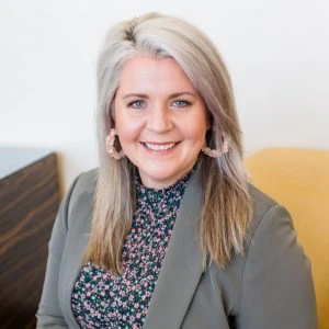 A woman with long, light gray-blond hair wearing a gray blazer and floral top sits on a yellow chair, smiling at the camera.
