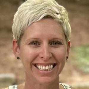 A person with short, light blonde hair smiling at the camera, wearing a sleeveless top and small earrings, with an outdoor background.