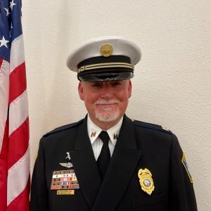 A uniformed fire department officer stands in front of a US flag and a beige wall, wearing a white peaked cap and displaying several badges and ribbons on his jacket.
