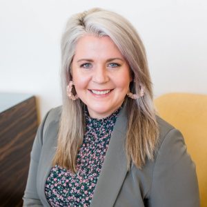 A woman with long, light gray hair wearing a gray blazer and floral blouse sits smiling on a yellow chair, embodying the welcoming spirit of BCEN conference speakers.