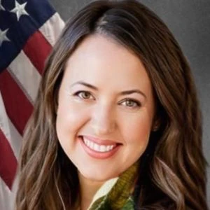 A woman with long brown hair smiles in front of a U.S. flag and a gray background.