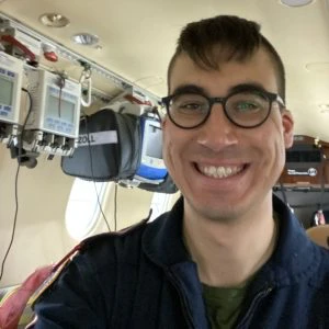 Person wearing glasses and a dark jacket smiling inside an aircraft, with medical equipment attached to the wall in the background.