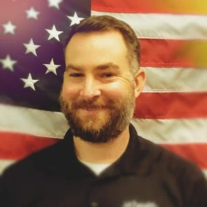 A man with a beard smiles in front of a blurred American flag backdrop, wearing a black collared shirt.