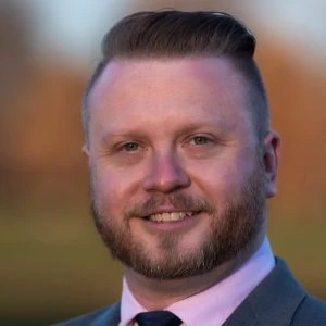 A man with light skin, short styled hair, and a trimmed beard is wearing a suit and tie, smiling at the camera outdoors.
