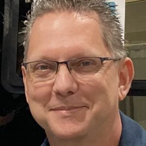 A middle-aged man with short, light brown hair and glasses smiles at the camera. He is indoors and wearing a dark shirt.