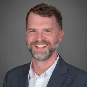 A man with short brown hair and a beard, wearing a white shirt and gray blazer, smiling at the camera against a gray background.