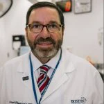 A man wearing glasses and a white lab coat with a striped tie stands in a medical or laboratory setting, facing the camera.