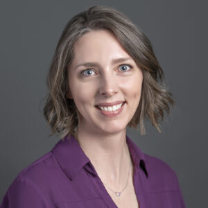 A woman with short, wavy brown hair and blue eyes is smiling at the camera, wearing a purple blouse and a necklace—one of the notable BCEN conference speakers—against a gray background.