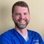 A man with light brown hair and a beard smiles at the camera while wearing a blue medical scrub top, standing in front of a beige background.