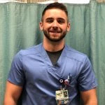 A man wearing blue medical scrubs and an ID badge stands and smiles in front of a teal curtain.