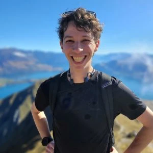 Person standing outdoors on a sunny day, smiling at the camera, with mountains and a body of water visible in the background.