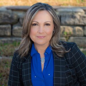 A woman with shoulder-length light brown hair wearing a blue blouse and a black checkered blazer sits outdoors in front of a stone wall, resembling one of the BCEN conference speakers.